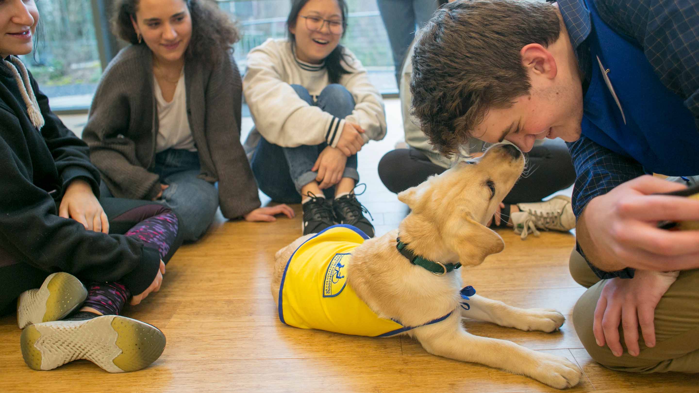 Students sitting around a puppy. One student is getting his face licked by a puppy.