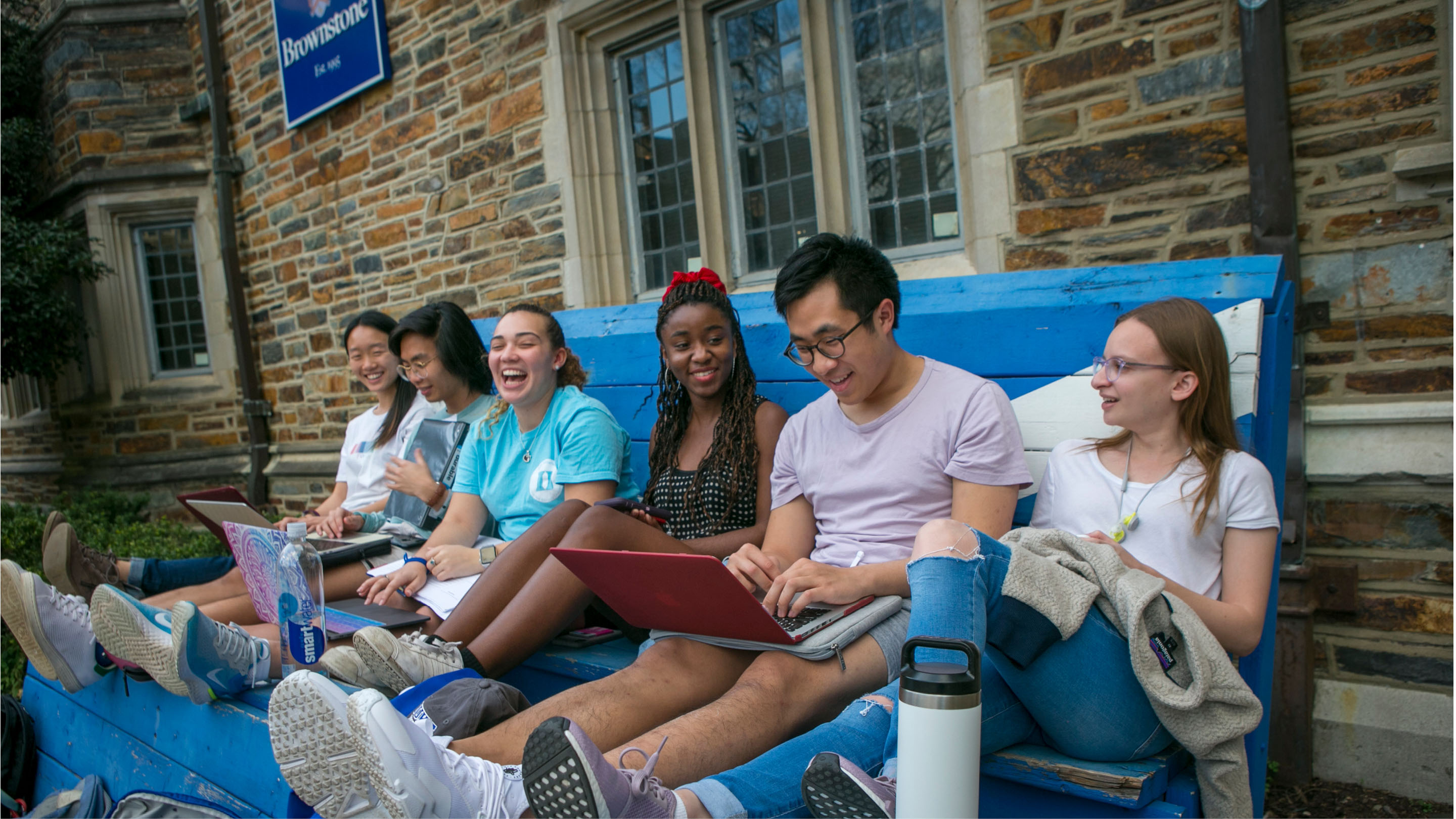 Students sitting together on oversized blue bench. Laughter and school work going on.