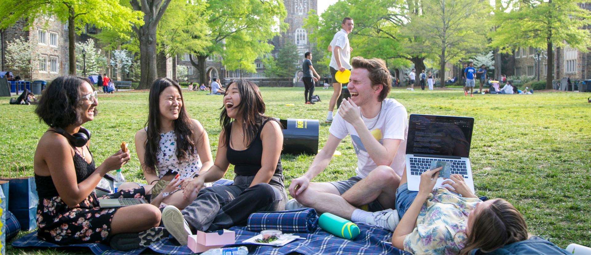 Four Duke students picnicking on a blanket on the campus Quad