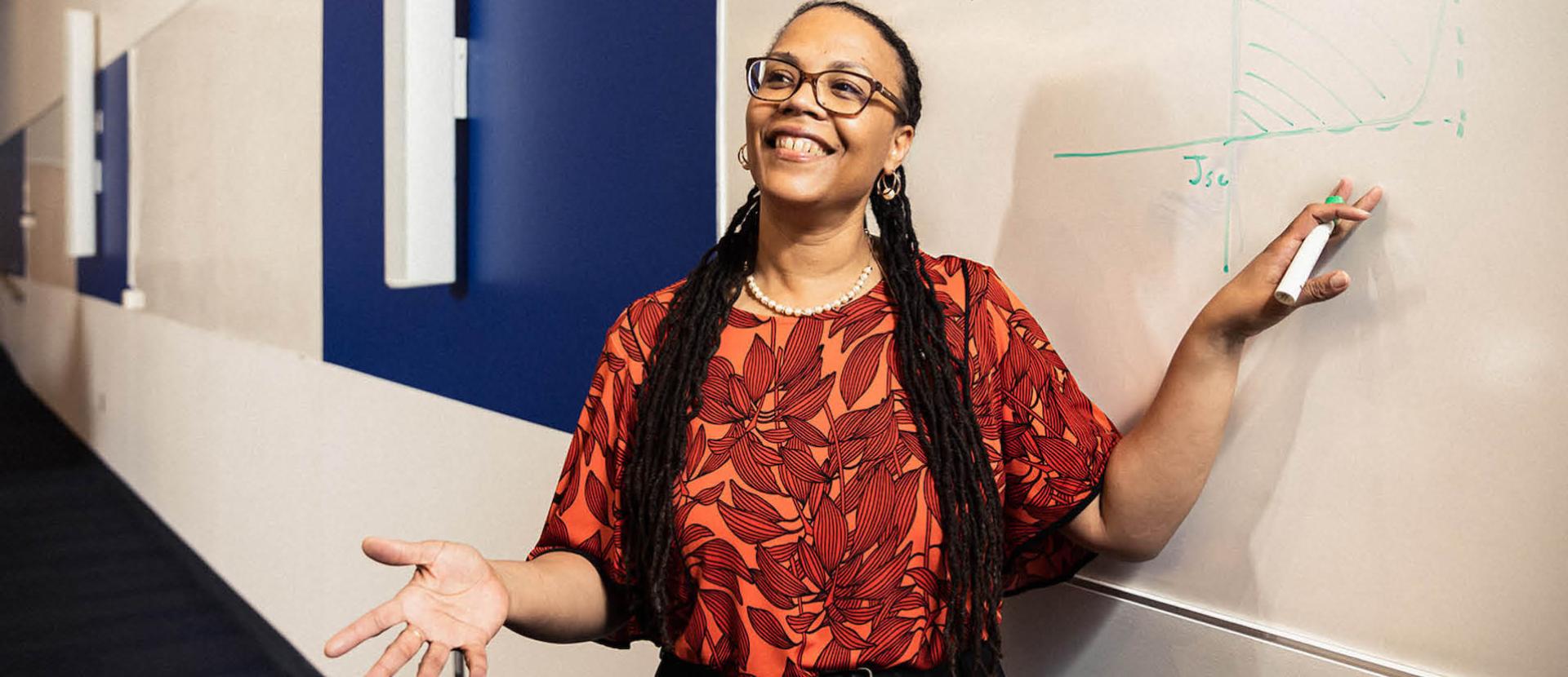 Adrienne-Stiff Roberts standing in front of a whiteboard smiling. Her right hand is outstretched, like she is explaining something. 