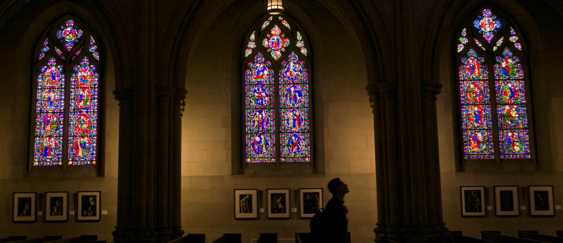 Student in shadows of Duke Chapel with three arched stained glass windows in background.