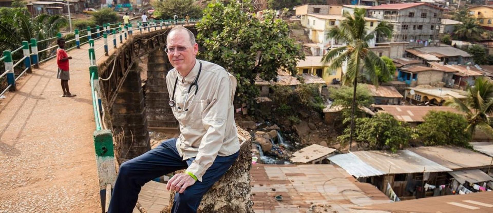The late Dr. Paul Farmer sits on a low wall in an unidenfitied location.
