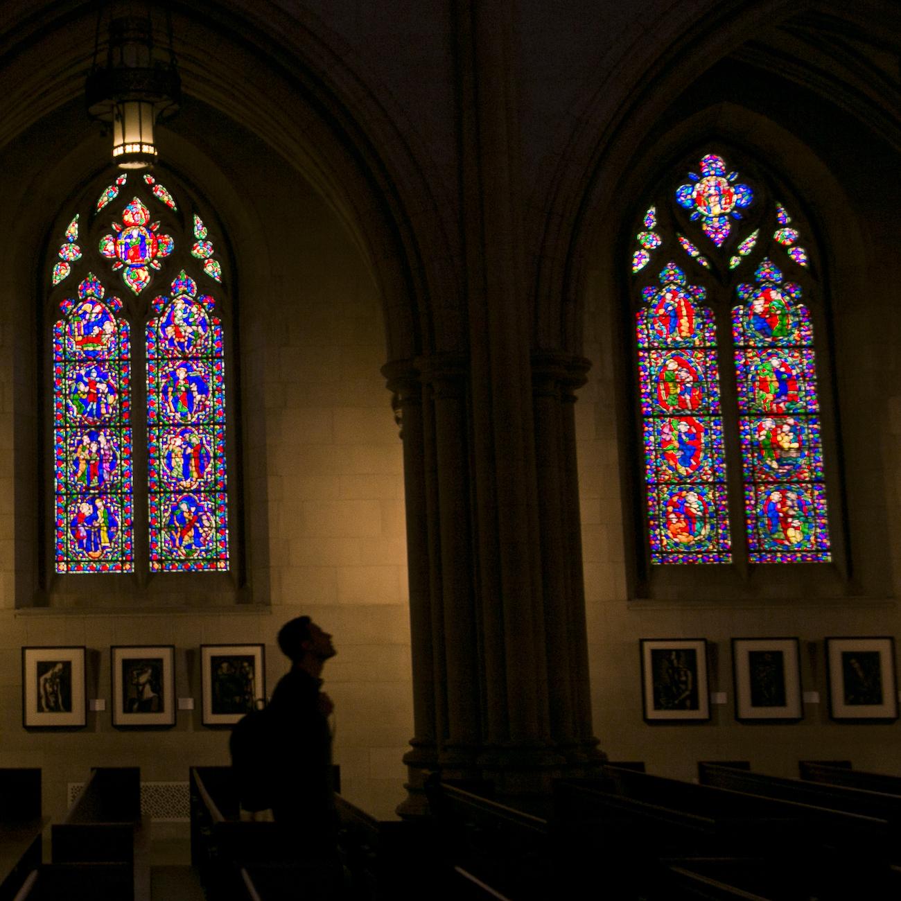 Student in shadows of Duke Chapel with three arched stained glass windows in background.