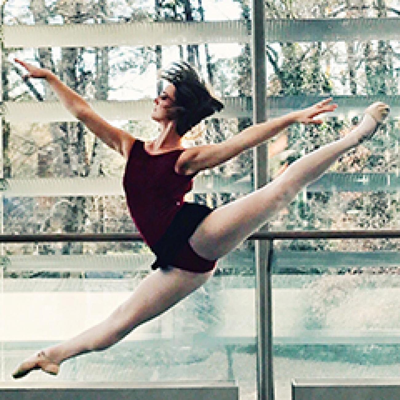 Student dancer doing split in the air in room with trees in the background